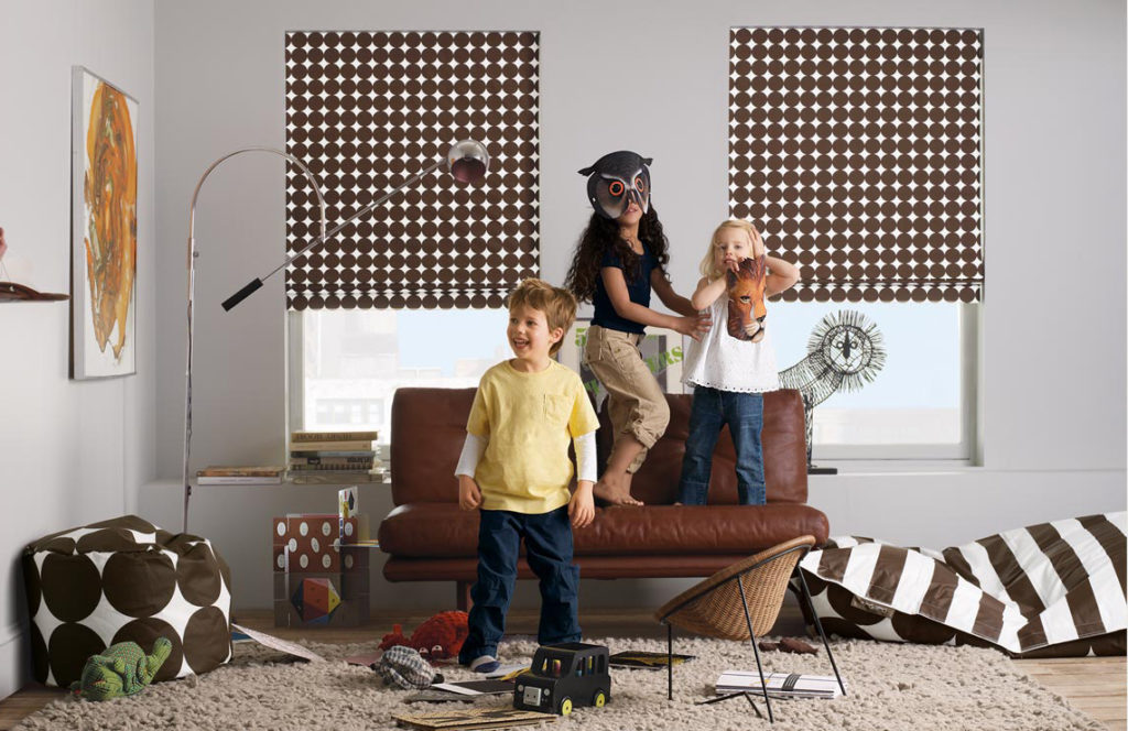 Three kids playing dress-up in a playroom with polka dot Flat Roman Shades on tall windows