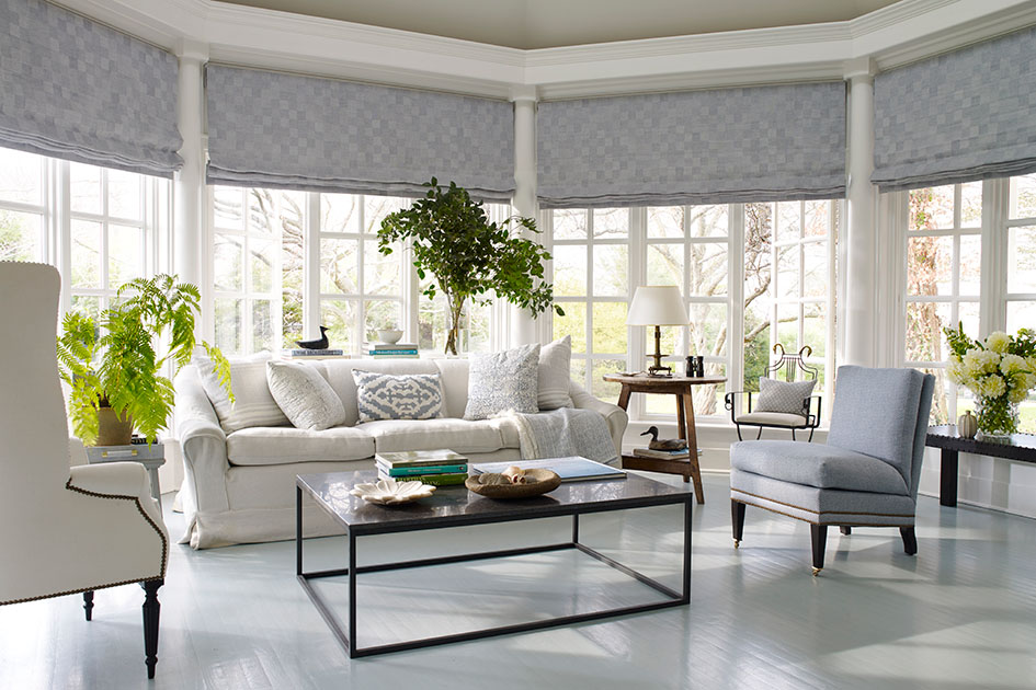 A large living room with a cool color scheme features flat roman shades which are one of the types of window shades