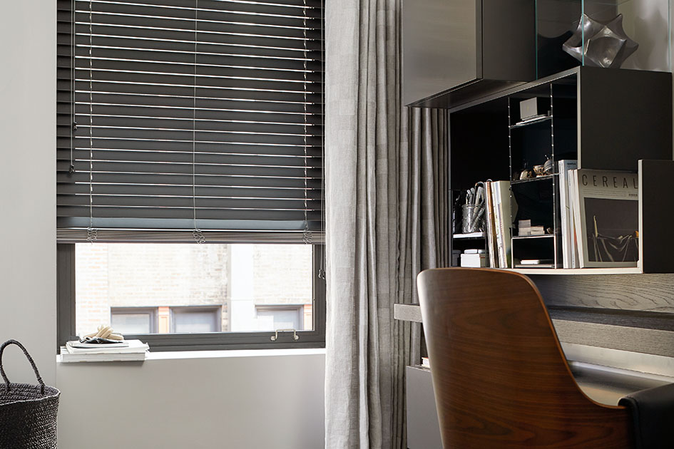 An office in a loft features a tall window with dark wood blinds which are one of the types of shades for windows