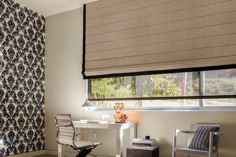 Large outside mounted Roman Shade in a home office with neutral fabric, black trim, and a wide window above a desk.