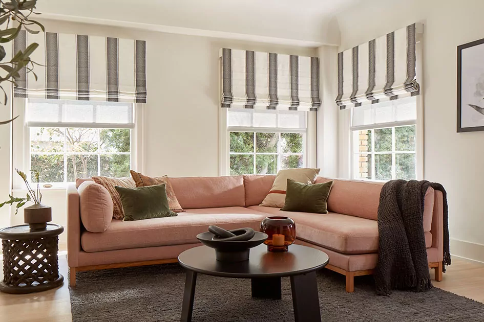 A casual living room with outside mount flat roman shades shows the difference between inside vs outside mount blinds