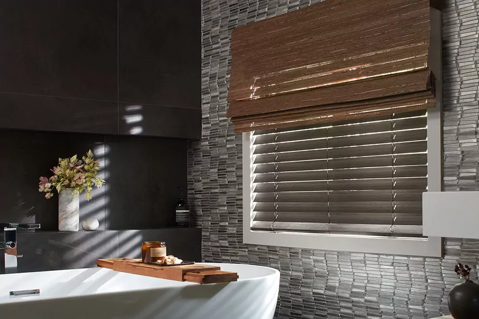 A dark bathroom has a small window with dark wood blinds and a woven wood shade showing how do cordless blinds work