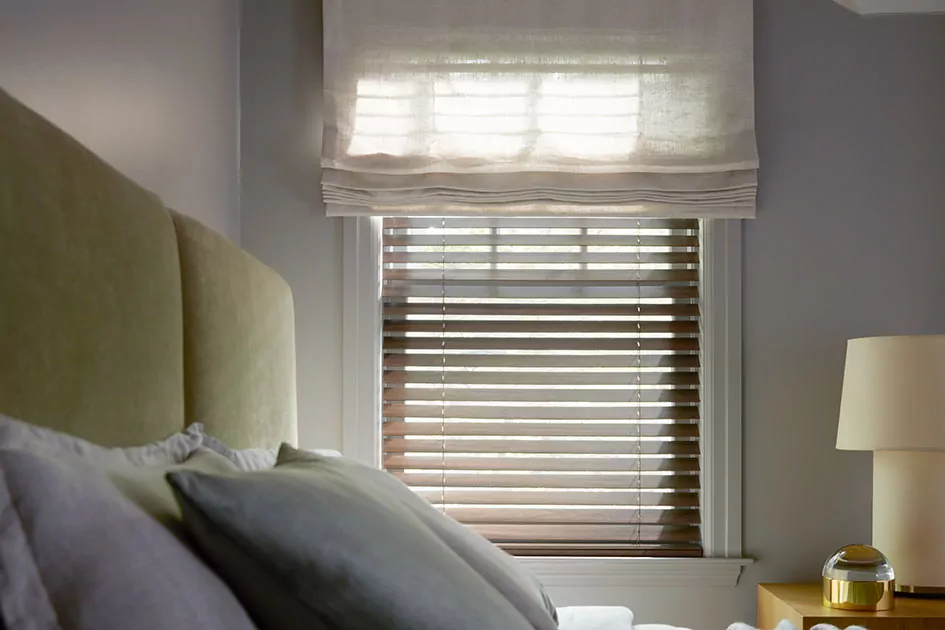 A bedroom has a tall window with wood blinds and flat roman shades, showing how do cordless blinds work when layered