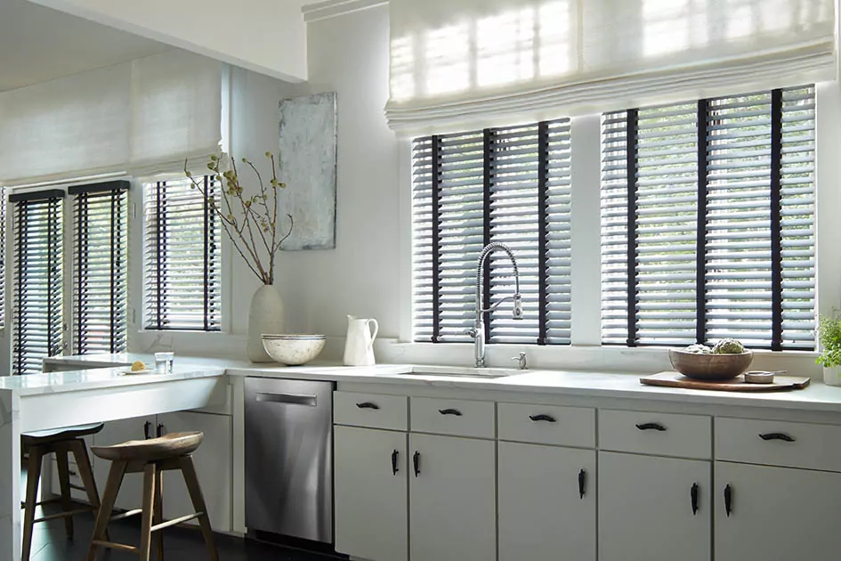 A modern kitchen features windows with wood blinds and flat roman shades showing how cordless blinds work when layered