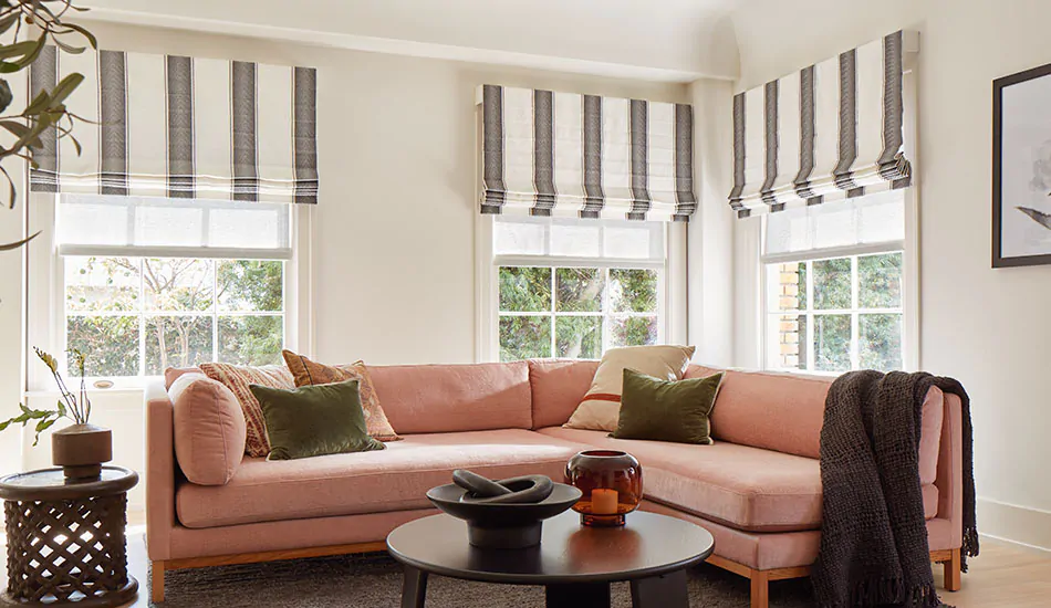 A casual family room with a coral couch has outside mount Cascade Roman Shades made of Nomad Stripe in Graphite