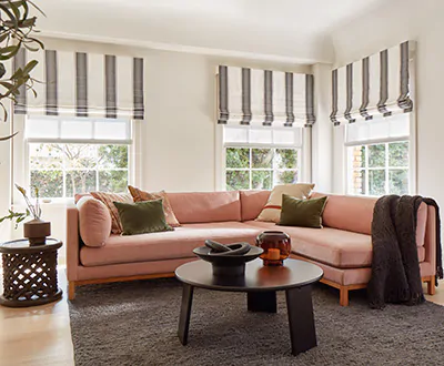 A family room with a rose-colored sectional couch benefits from three windows with cascade roman shade treatments