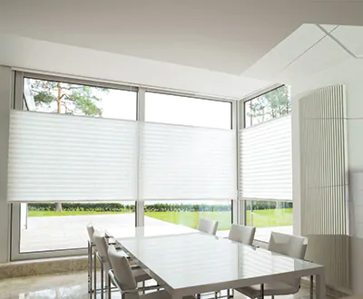 A minimalist dining room with white walls and a white table has corner windows with Cellular Shades made of Avalon in White