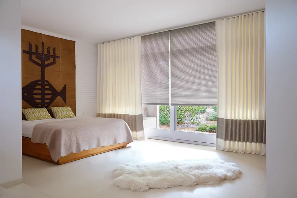 A bedroom features cream colored ripple fold drapery layered over cellular shades for sliding glass doors