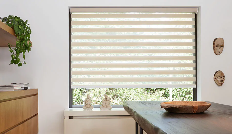 A dining area with Buddha statues on the window sill and a rustic wood table has Zebra Blinds made of Windansea in Canyon