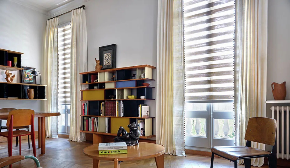 A lounge with wood herringbone floors has zebra blinds made of Windansea in Canyon layered with Raw Silk drapes in Glacier