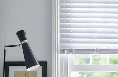 Faux Wood Blinds made of 2-inch Blanc material match the white walls in a room with a black desk lamp