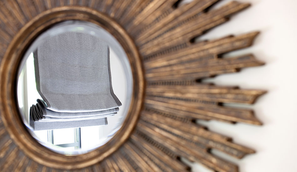 A mirror with a sunburst design reflects light from a window partially covered with a Flat Roman Shade of Baldwin in Stone