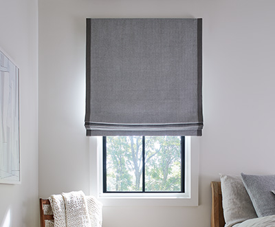 A Cordless Roman Shade made of Baldwin in Stone in a cozy bedroom shows one style of pull down window shades