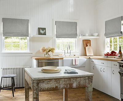 A farmhouse kitchen features a rustic table and butcher block countertop and Flat Roman Shades made of Cotton in Chino