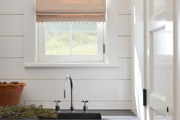 Transitional window treatments include a Flat Roman Shade in Denim, Sand, above a black utility sink in a white room