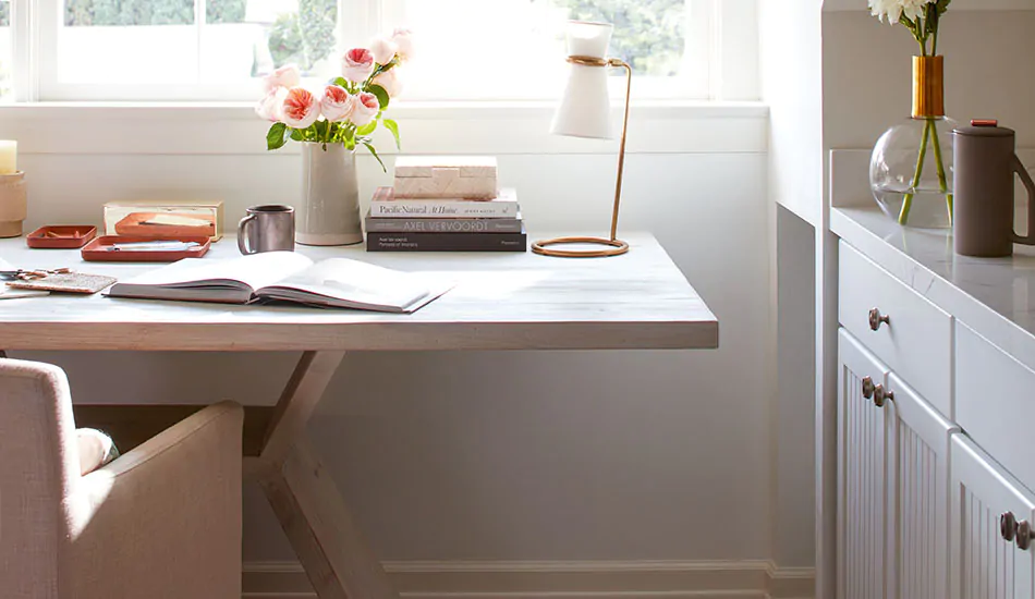 An office features simple, streamlined furniture in light colors including a whitewashed wood table and pink chair