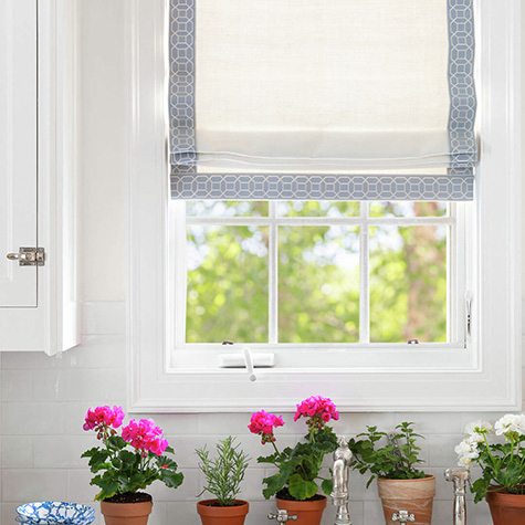 Small potted flowers sit below Roman shades for kitchen windows made of Luxe Linen in Oyster with Sakiori Embroidered trim