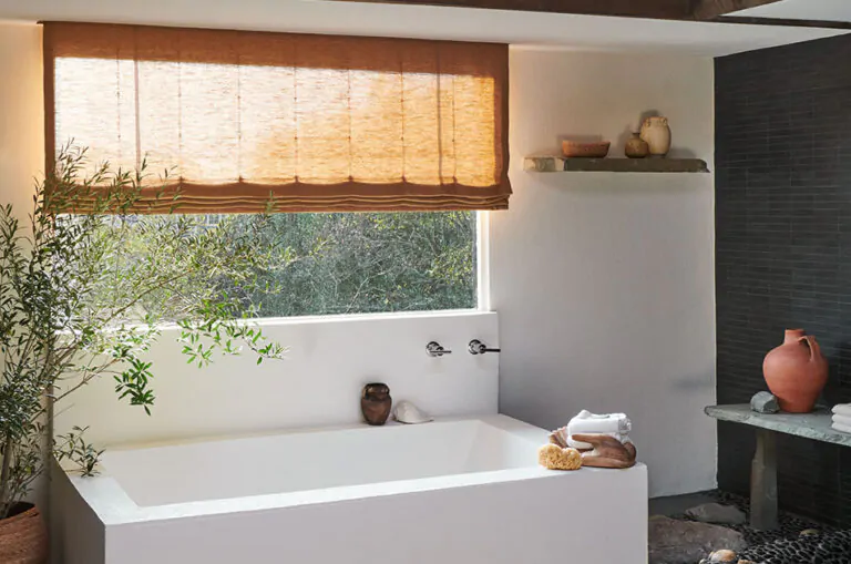 A Flat Roman Shade made of Lisbon Woven in Bronze provides bathroom window privacy in a tranquil bathroom with stone decor