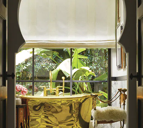 A Flat Roman Shade made of Nomad Stripe in Blanco offers bathroom window privacy in a luxe bathroom with a gold tub