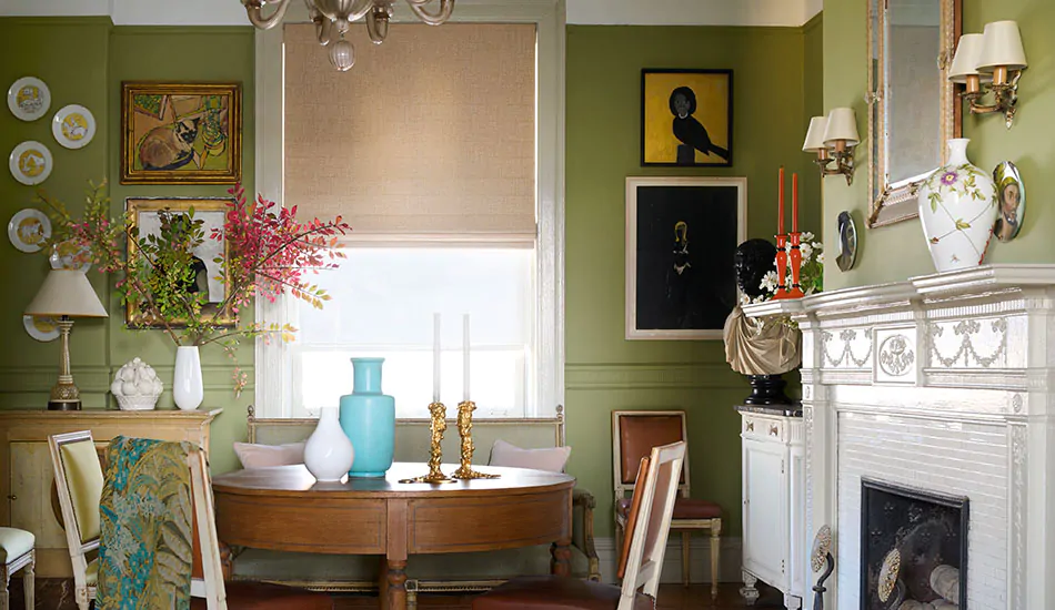 A dining room with green walls has flat roman shades made of Aerial in Tan and a Solar Shade of 3% Thermo in cloud