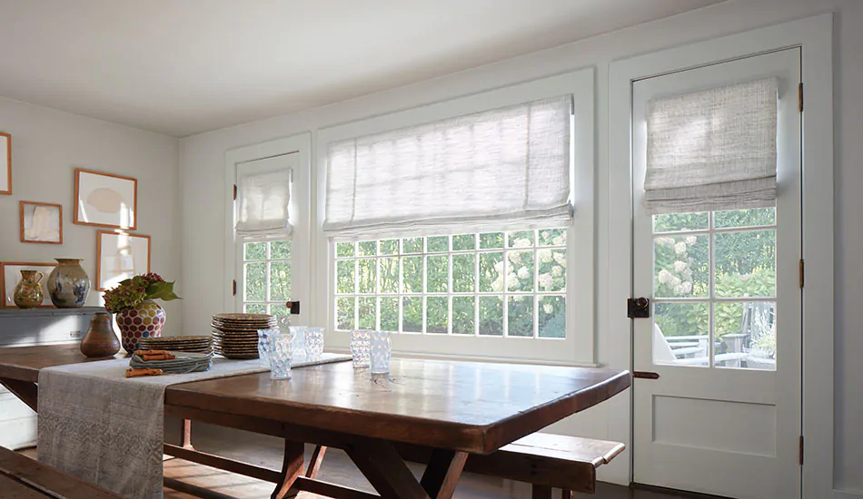 A bright dining room with a long wood table features white Roman Shades, showing the difference between blinds vs shades