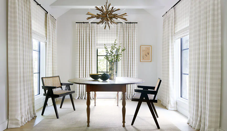 A dining room has layered window treatments, Tailored Pleat Drapery and Flat Roman Shades both made of Emerson in Shea