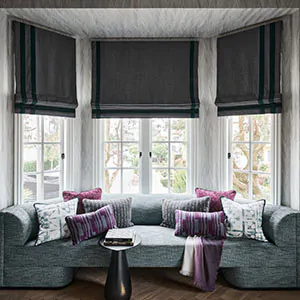 Window treatments for bay windows include Flat Roman Shades made of C.O.M. in a sitting area with a dark grey color scheme