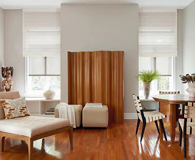 A living room with wood flooring and a table is well lit by two vertical windows in the flat roman shade style