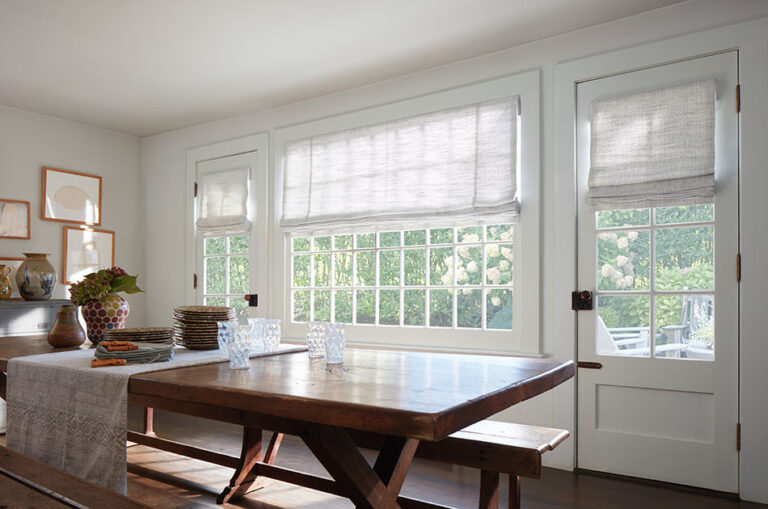 A bright white kitchen with a rustic wood table features a door window shade made of a Roman Shade in Shoreline, Cloud