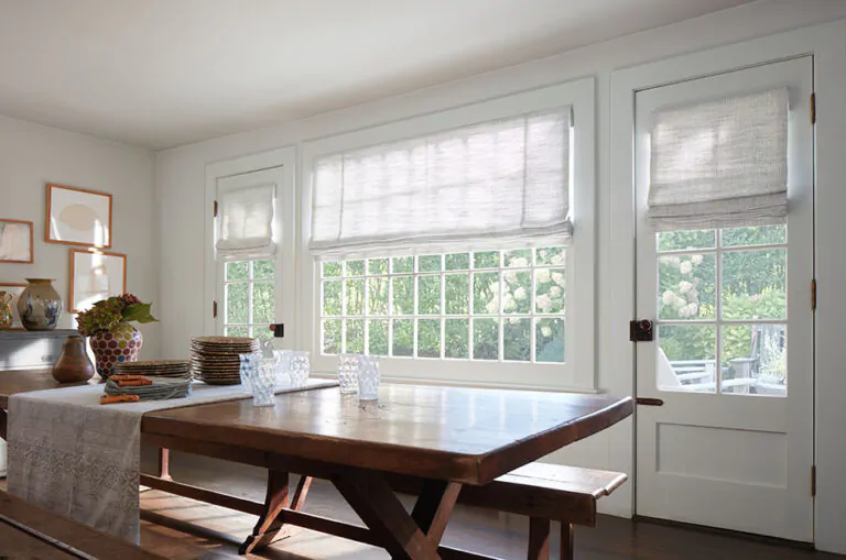 A bright white kitchen with a rustic wood table features a door window shade made of a Roman Shade in Shoreline, Cloud