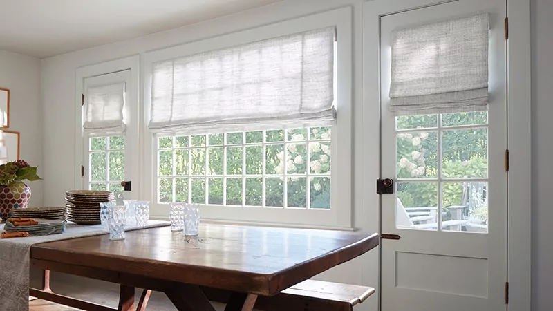 A large dining room features wide, bright windows with light flat roman shades that contrast with a dark wood table