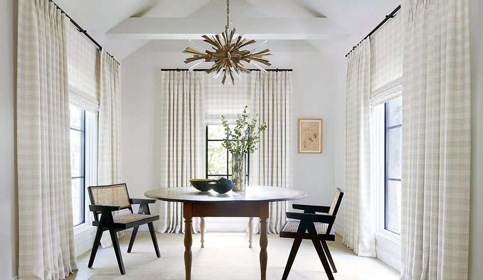A transitional dining room has a round wood table and Flat Roman Shades and Tailored Pleat Drapery made of Emerson in Shea