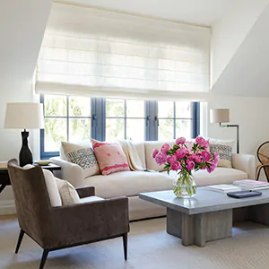 A sitting room with pink flowers and white light filtering Roman Shades shows the difference between blinds vs shades