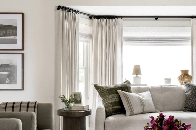 A living room features flat roman shades and tailored pleat drapery made of linen sheers in oatmeal as bay window curtains