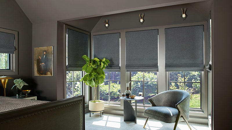 A bedroom with warm dark grey walls features tall windows with Flat Roman Shades made of Wool Blend in Charcoal