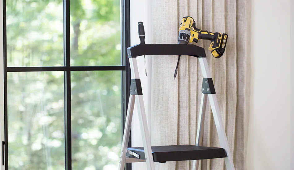 A step ladder with a drill resting on the top stands next to a window with drapery on it