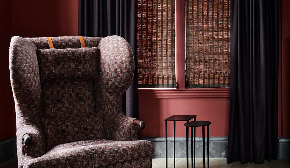 A sitting room has bold dark red walls a Inverted Pleat Drapery made of Wool Blend in Charcoal layered with a woven shade