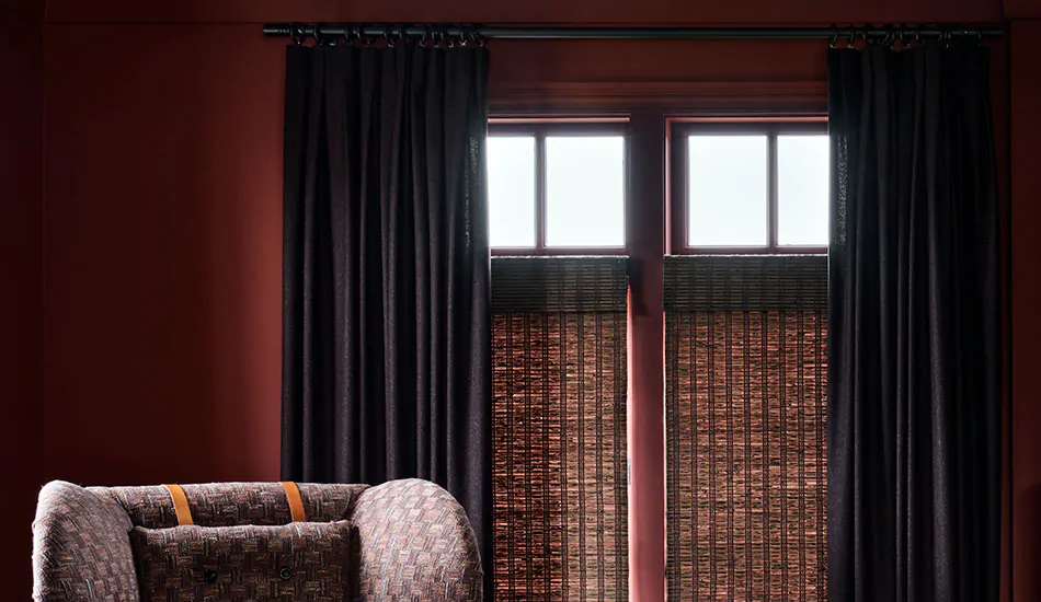 A sitting room has bold dark red walls a Inverted Pleat Drapery made of Wool Blend in Charcoal layered with a woven shade