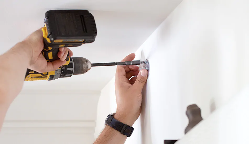 A close up shows a professional with a drill and bracket showing one step for how to hang curtains