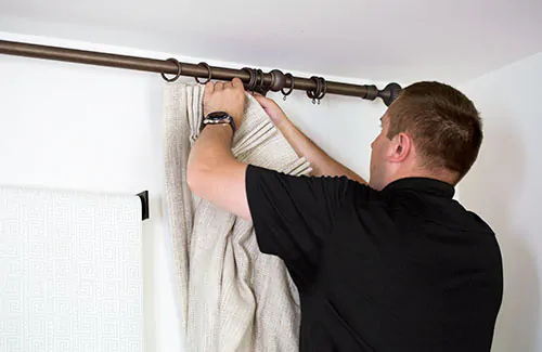 A professional installer attaches Pinch Pleat Drapery made of Shoreline in Reed on a curtain rod with rings