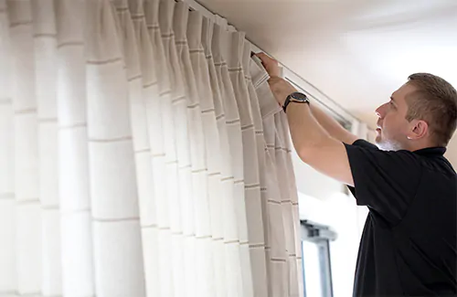 A professional installer attaches Pinch Pleat Drapery made of Tidal Line in Sand on a track system with hooks