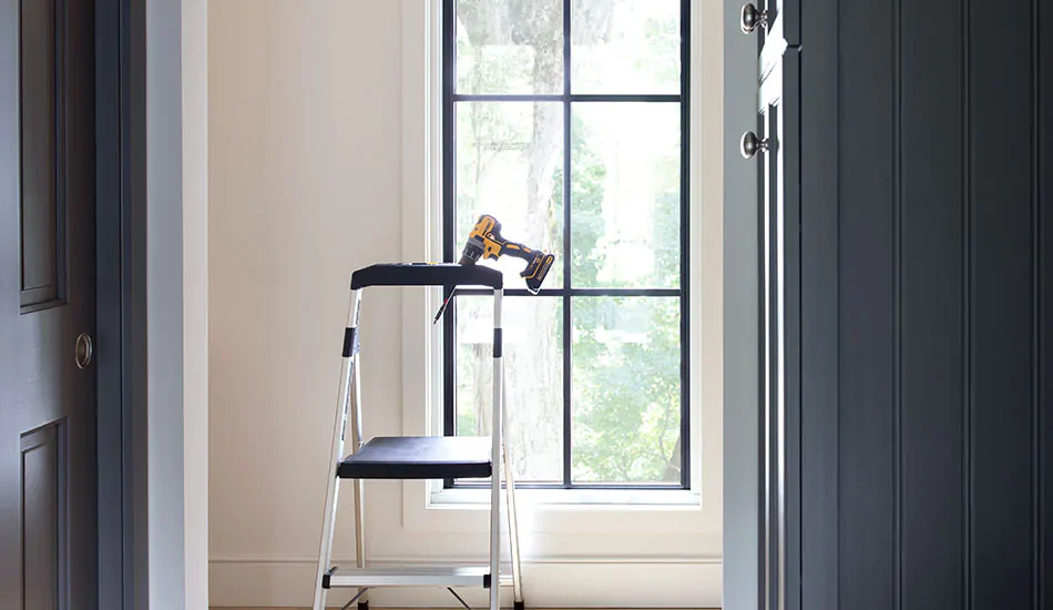 A step ladder with a drill sits in front of a bare window with no window treatments in a bright, small room