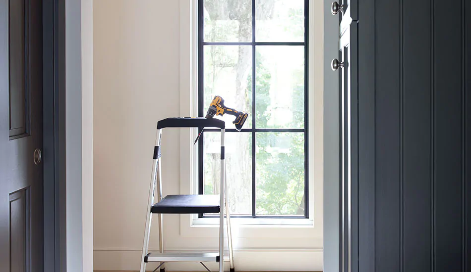 A step ladder with a power drill on top sit in front of a window in preparation for window treatment installation