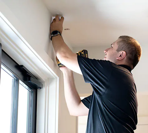 A professional installer standing on a step ladder smiles as he marks where to place a bracket beside a window
