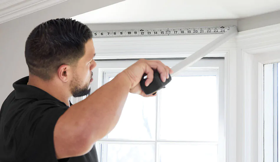 A professional window treatment installer measures a side window in a bay window with a silver measuring tape