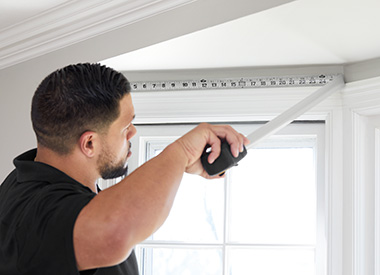 A professional window treatment installer measures the width of a window with a metal tape measure