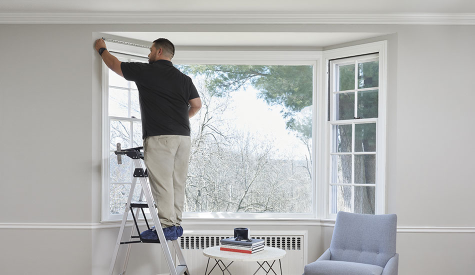 A window treatment installation professional stands on a step ladder and measures a bay window for Roman Shade installation