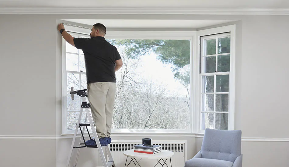 A window treatment installation professional stands on a step ladder and measures a bay window for Roman Shade installation
