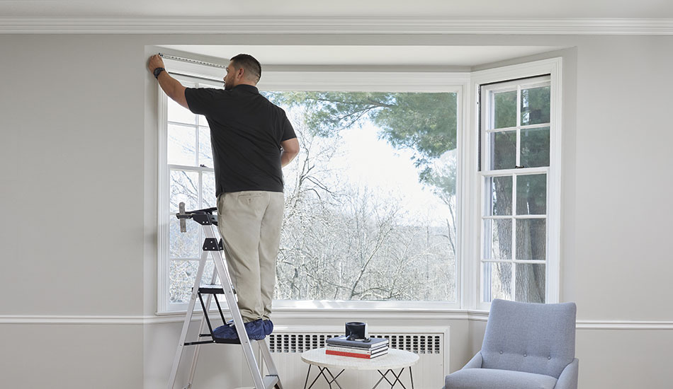 A professional window treatment installer stands on a step ladder at a bay window with a tape measure