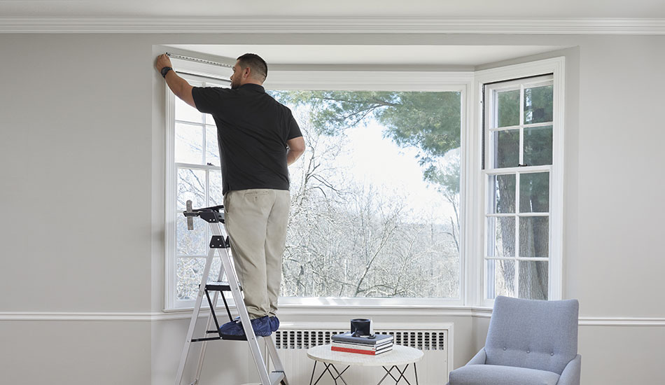A professional window treatment installer measures window in a bay window in preparation for installation of woven shades